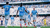 Manchester City players celebrating a goal at the Etihad Stadium during a match, with a crowd of spectators in the background.