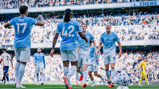 Manchester City players celebrating a goal at the Etihad Stadium during a match, with a crowd of spectators in the background.