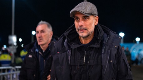 Two individuals are walking outside at night wearing heavy jackets; background includes people in safety vests and bright lights.
