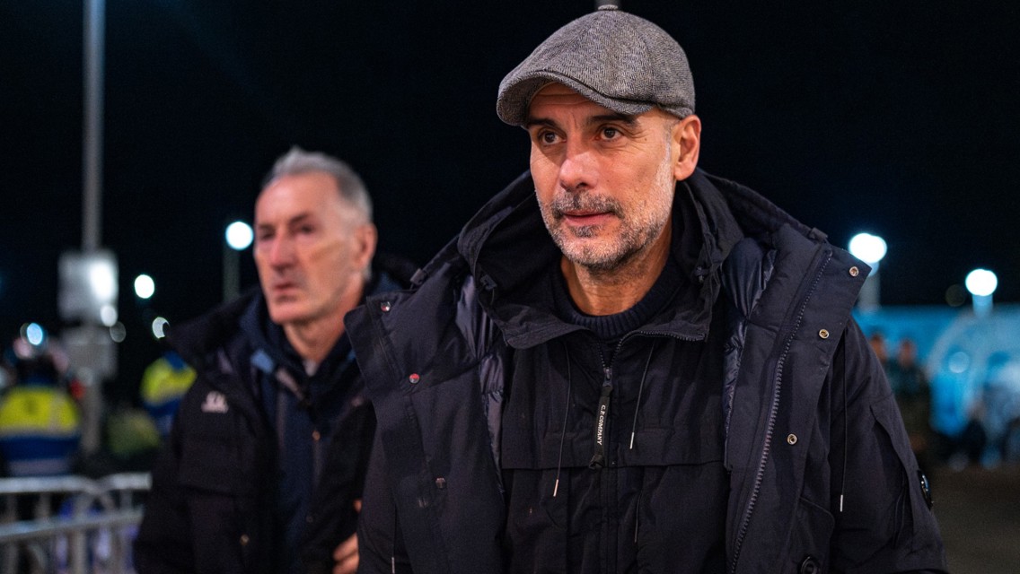 Two individuals are walking outside at night wearing heavy jackets; background includes people in safety vests and bright lights.
