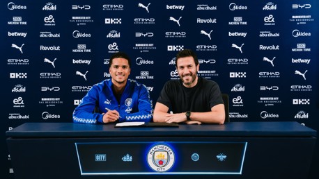 Two individuals sitting at a table with Manchester City logos and sponsors in the background, appearing to sign a contract.