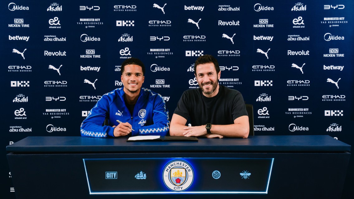 Two individuals sitting at a table with Manchester City logos and sponsors in the background, appearing to sign a contract.