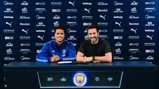 Two individuals sitting at a table with Manchester City logos and sponsors in the background, appearing to sign a contract.
