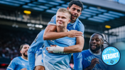 Football players wearing Manchester City jerseys celebrate on the field. One player is giving a piggyback ride to another while others join in the celebration.