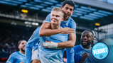 Football players wearing Manchester City jerseys celebrate on the field. One player is giving a piggyback ride to another while others join in the celebration.