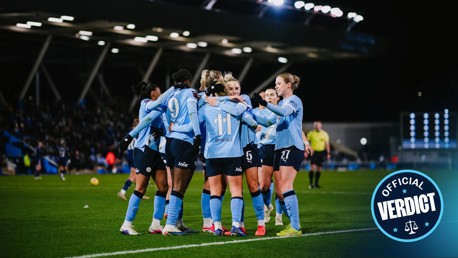 Women's soccer team with blurred faces celebrating on the field, wearing light blue jerseys with numbers and sponsor logos.