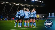 Women's soccer team with blurred faces celebrating on the field, wearing light blue jerseys with numbers and sponsor logos.
