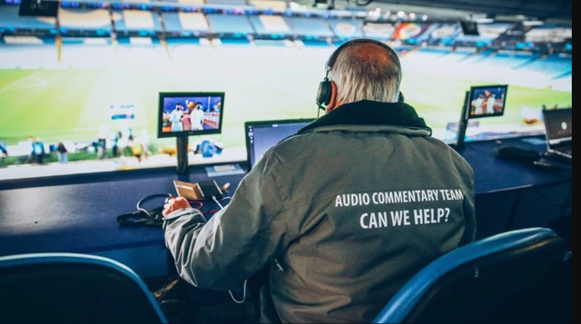 An audio commentary team member is seated at a workspace overlooking a stadium field, with monitors and equipment on the desk, wearing a jacket labeled 'AUDIO COMMENTARY TEAM CAN WE HELP?'