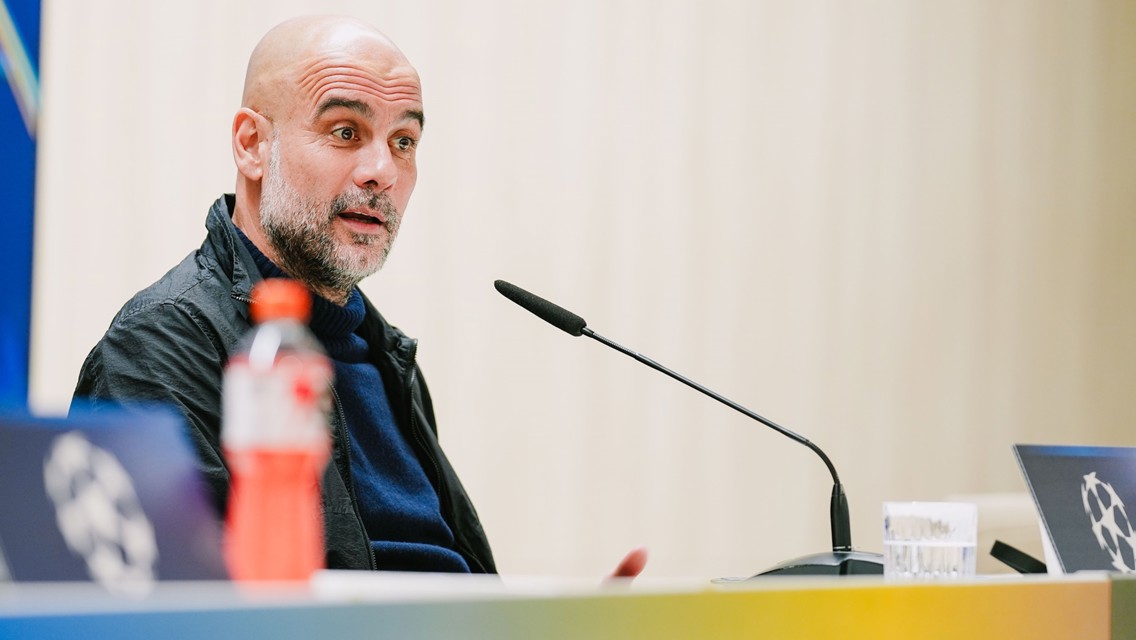 Person at a press conference with a microphone on the table, blurred face, wearing a jacket.
