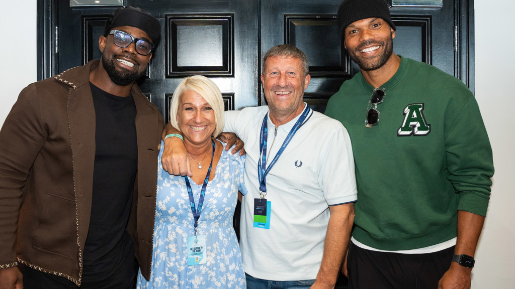 Four people are smiling and posing in front of a black door, wearing casual clothes with lanyards.