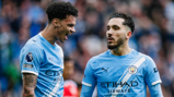 Two Manchester City players wearing the team's blue kit during a match, featuring the Premier League logo and sponsor branding on their shirts.