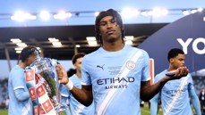 A Manchester City youth player holds the FA Youth Cup trophy on the field, wearing a team jersey with Etihad Airways sponsorship.