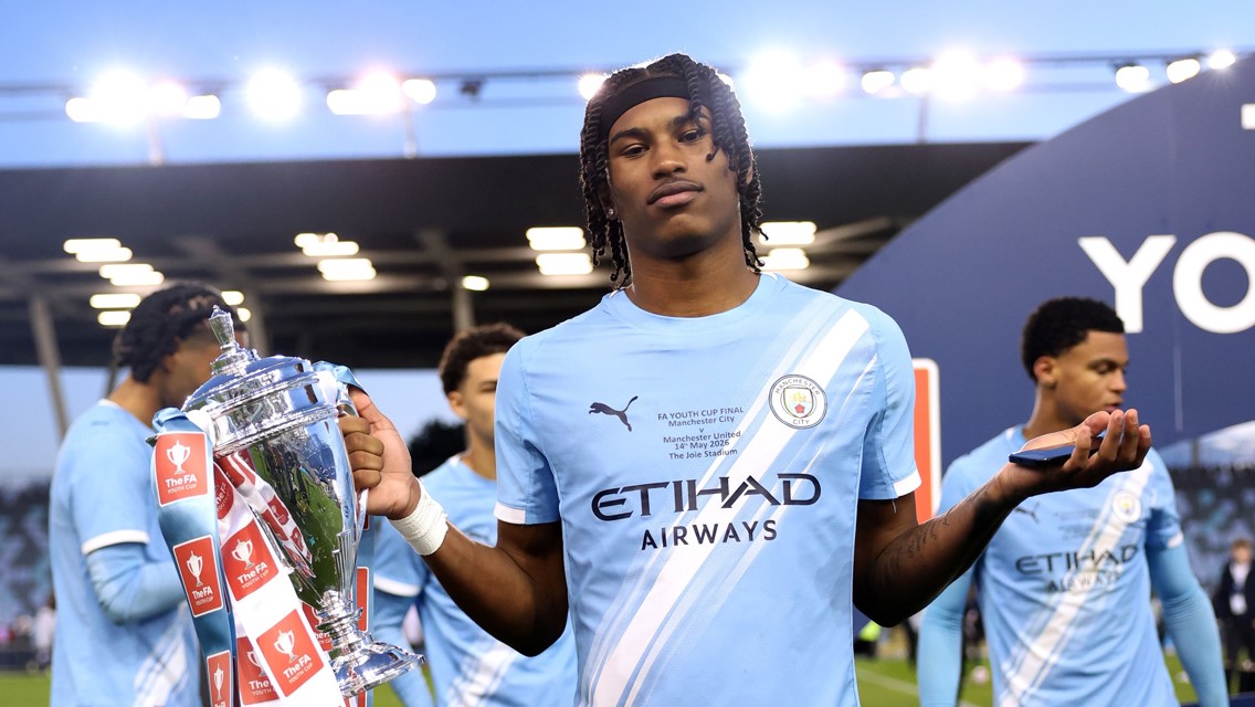 A Manchester City youth player holds the FA Youth Cup trophy on the field, wearing a team jersey with Etihad Airways sponsorship.