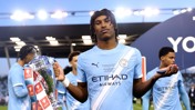 A Manchester City youth player holds the FA Youth Cup trophy on the field, wearing a team jersey with Etihad Airways sponsorship.