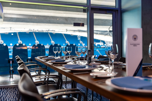 A dining table setup inside a stadium hospitality area with a view of the field. Glasses, plates, and menus are on the table. Blue stadium seats visible outside.