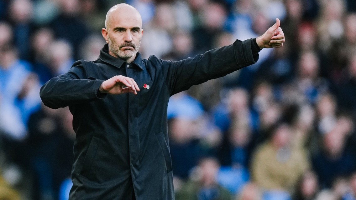 A man with a bald head wearing a black jacket gives a thumbs up gesture in a crowded setting.