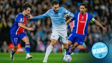 A Manchester City player in a blue kit is trying to dribble past two Paris Saint-Germain players in a red and blue kit during a football match.