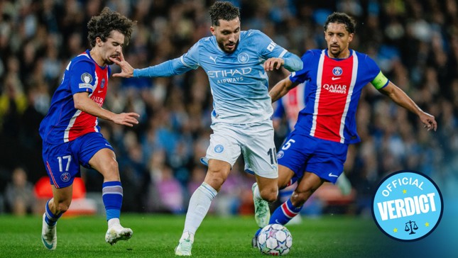 A Manchester City player in a blue kit is trying to dribble past two Paris Saint-Germain players in a red and blue kit during a football match.