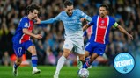 A Manchester City player in a blue kit is trying to dribble past two Paris Saint-Germain players in a red and blue kit during a football match.