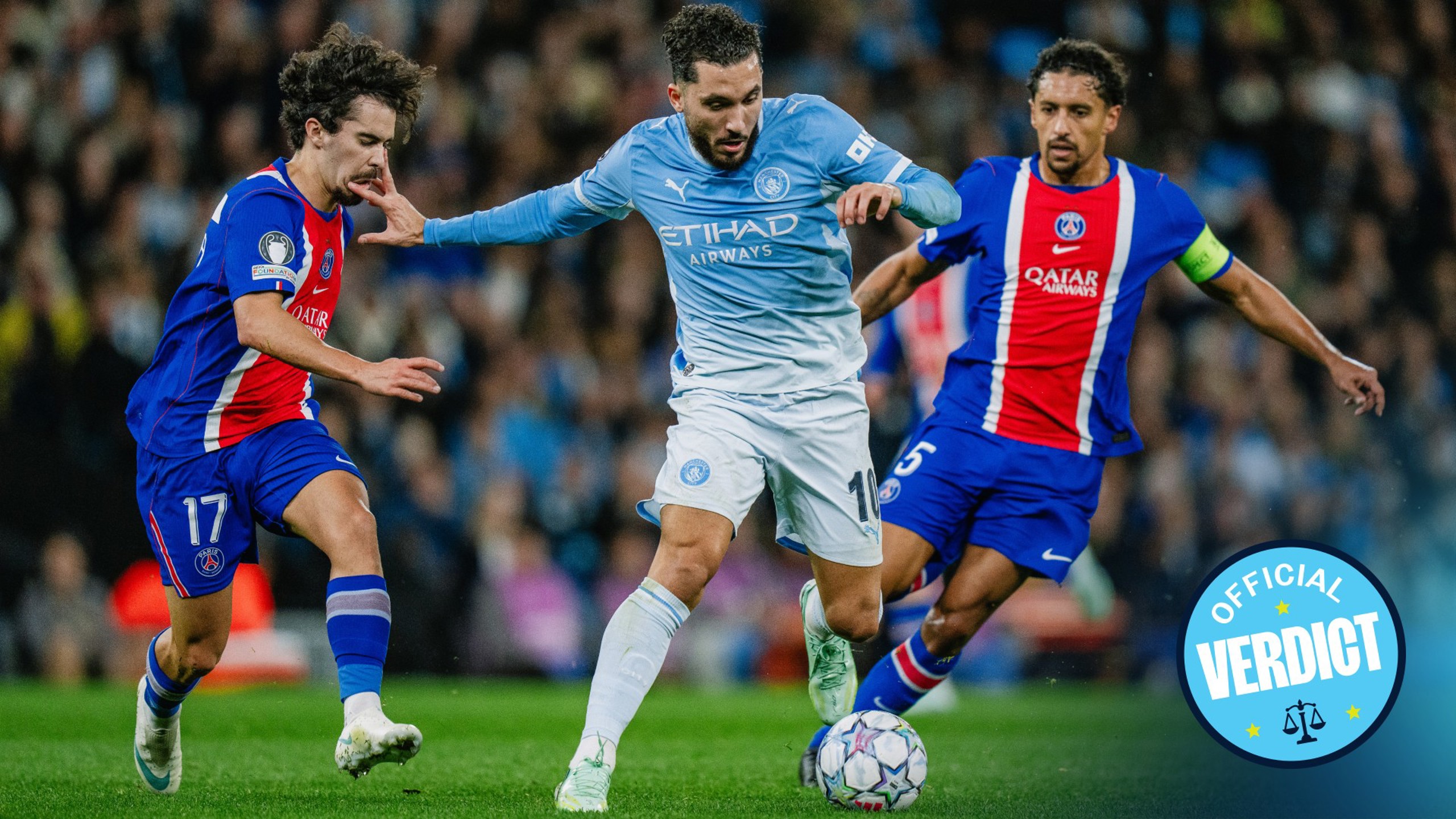 A Manchester City player in a blue kit is trying to dribble past two Paris Saint-Germain players in a red and blue kit during a football match.