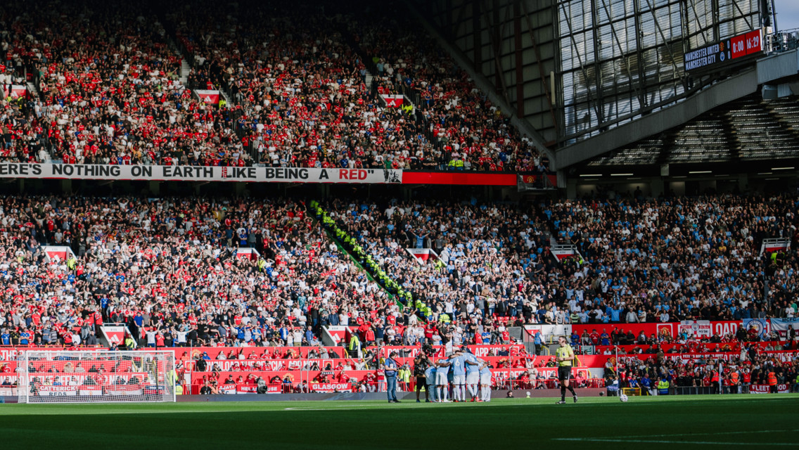 Football match at Old Trafford between Manchester United and Manchester City with a crowded stadium and fans wearing club colors.