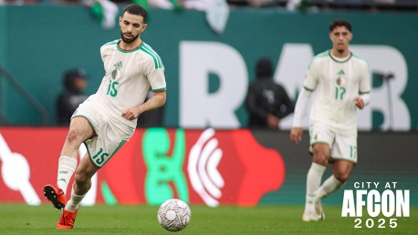 Two soccer players in white uniforms with green accents are playing on the field during AFCON 2025. The player in front has the number 15 on his jersey and is actively controlling the ball.