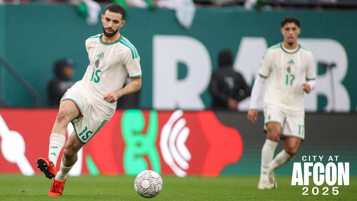 Two soccer players in white uniforms with green accents are playing on the field during AFCON 2025. The player in front has the number 15 on his jersey and is actively controlling the ball.