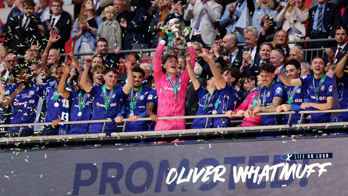 A sports team celebrates a trophy victory, with players wearing blue jerseys and a goalkeeper in pink holding the trophy aloft while confetti falls.