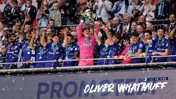 A sports team celebrates a trophy victory, with players wearing blue jerseys and a goalkeeper in pink holding the trophy aloft while confetti falls.