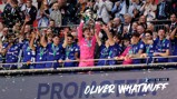 A sports team celebrates a trophy victory, with players wearing blue jerseys and a goalkeeper in pink holding the trophy aloft while confetti falls.