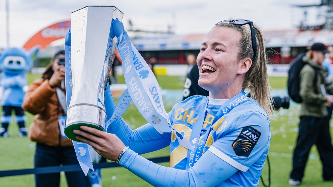 A football player in a blue jersey celebrates with a silver trophy on the field. The trophy has Barclays branding, and the player wears sunglasses and has a championship ribbon around the neck. In the background, a mascot and spectators are visible.