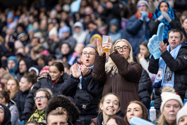 Crowd at a sporting event, people wearing warm clothing, some with Manchester City logos, a person holding a drink, foam finger visible.