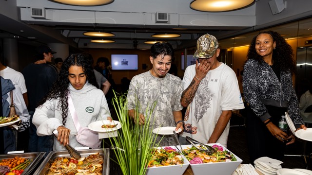 Several people are enjoying a buffet at a social gathering, selecting food from trays filled with various dishes. The atmosphere is casual and friendly.