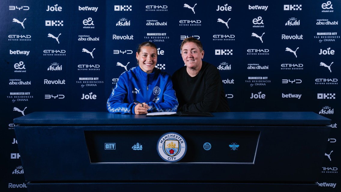 Two people at a table with Manchester City logos and sponsors, one in sports attire signing a document.