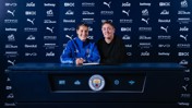Two people at a table with Manchester City logos and sponsors, one in sports attire signing a document.