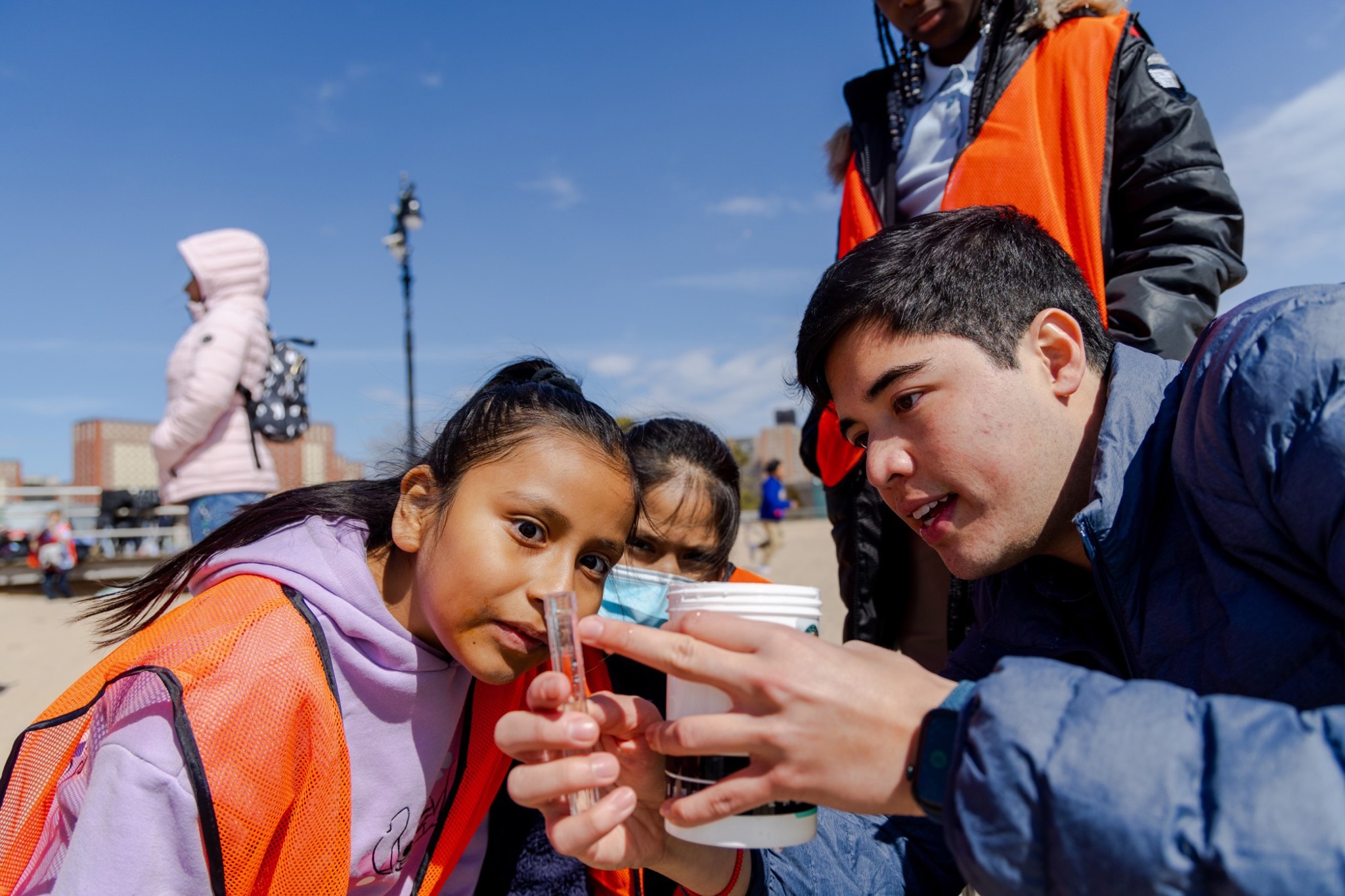A group of kids wearing orange vests is gathered around a person showing them something in a cup on a beach.