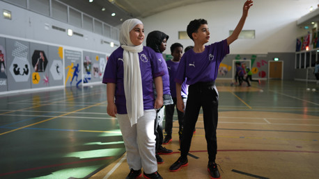 Children wearing purple sports jerseys in an indoor sports hall. They appear engaged in a group activity, with some looking towards the ceiling or gesturing. The sports hall is decorated with colorful athletic-themed murals.