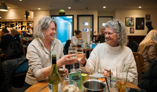 Two women sit at a table in a cafe or restaurant, smiling and toasting with glasses of wine. The table has drinks and a bottle of wine, and the venue is bustling with other patrons.