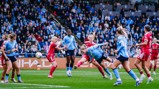 Women's soccer match with players in blue and red jerseys in action on the field, surrounded by a crowd of spectators.