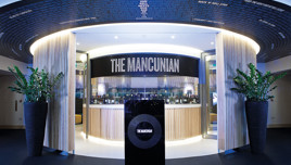 Entrance to The Mancunian restaurant with a modern design, featuring large potted plants and curved wooden panels. Restaurant name is prominently displayed above the entrance.