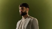 A man with a beard wearing a checkered blazer and a gray t-shirt, standing in profile against a green background.