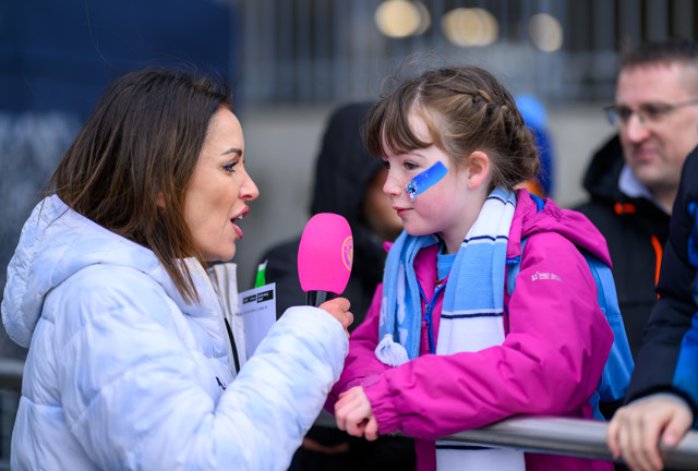 A young girl wearing a Manchester City scarf is being interviewed with a pink microphone, likely by a media representative at a stadium.