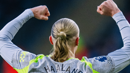 A football player with long hair and jersey labeled 'Haaland' celebrates by raising both fists. The jersey features Premier League patches and has a sponsor logo on the sleeves.