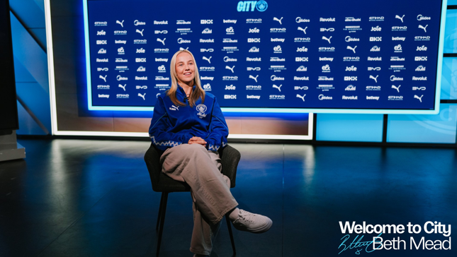 A person wearing a Manchester City jacket is sitting in front of a backdrop featuring sponsor logos. The text 'Welcome to City Beth Mead' is visible.