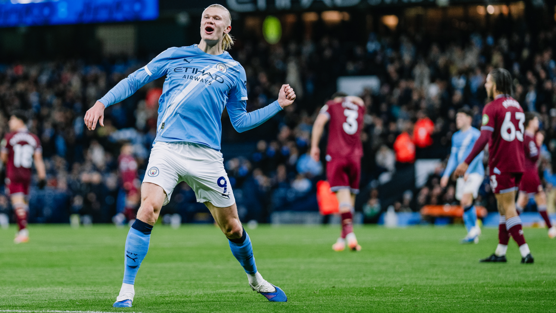 Soccer player in a Manchester City kit celebrating a goal on the field against a maroon-clad team, in front of a stadium crowd.