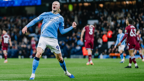 Soccer player in a Manchester City kit celebrating a goal on the field against a maroon-clad team, in front of a stadium crowd.