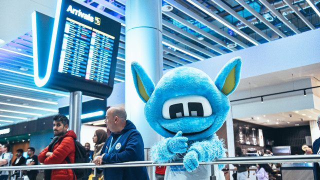 The Manchester City mascot Moonchester is seen at an airport, standing near the arrivals screen. It's surrounded by a crowd of people observing the arrivals screen.