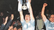 A football player in a blue and white striped jersey holds up a trophy with blue and white ribbons, surrounded by cheering crowd.