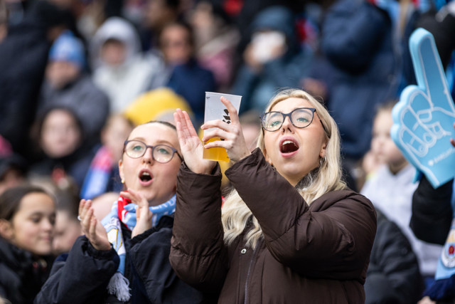 A crowd of people at an event, with one person holding a drink and taking a photo using a smartphone. Others are blurred in the background, holding up foam fingers and clapping.