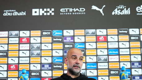 A person sitting at a press conference with a backdrop featuring various brand logos.